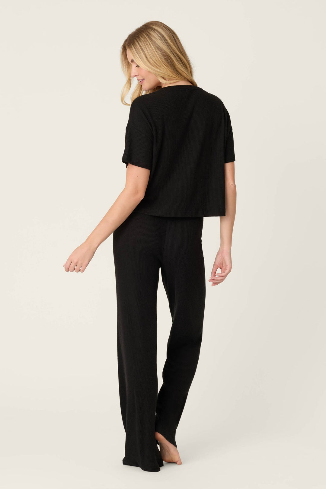 A woman wearing a black short-sleeve cropped top and matching black wide-leg pants, shown from the back against a plain light beige studio background.
