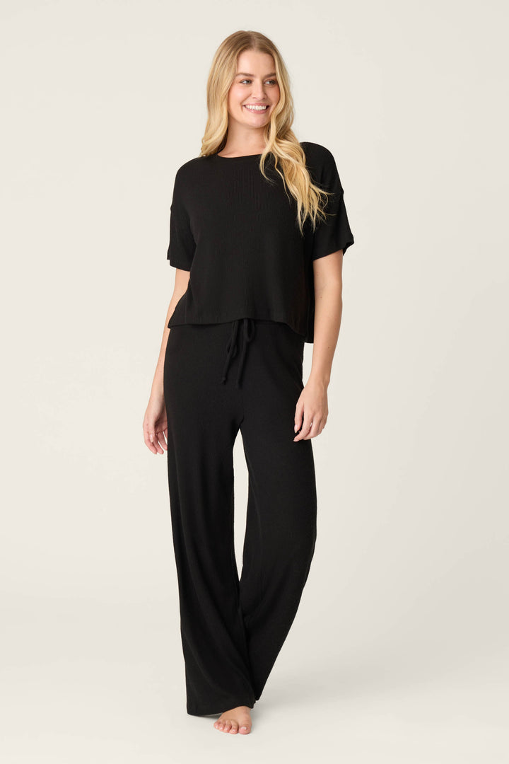 A woman modeling a black two-piece loungewear set consisting of a short-sleeve cropped top with a drawstring waist and matching wide-leg pants, photographed against a plain light gray studio background.