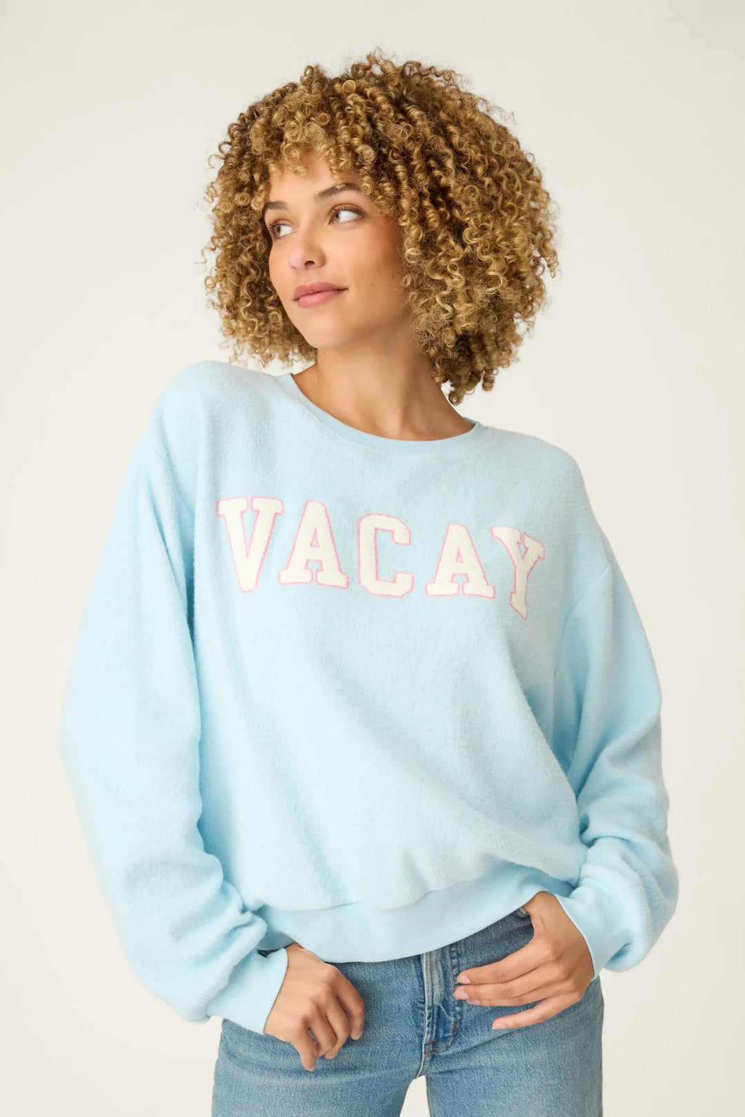 Woman wearing a light blue sweatshirt with 'VACAY' text, standing against a plain background