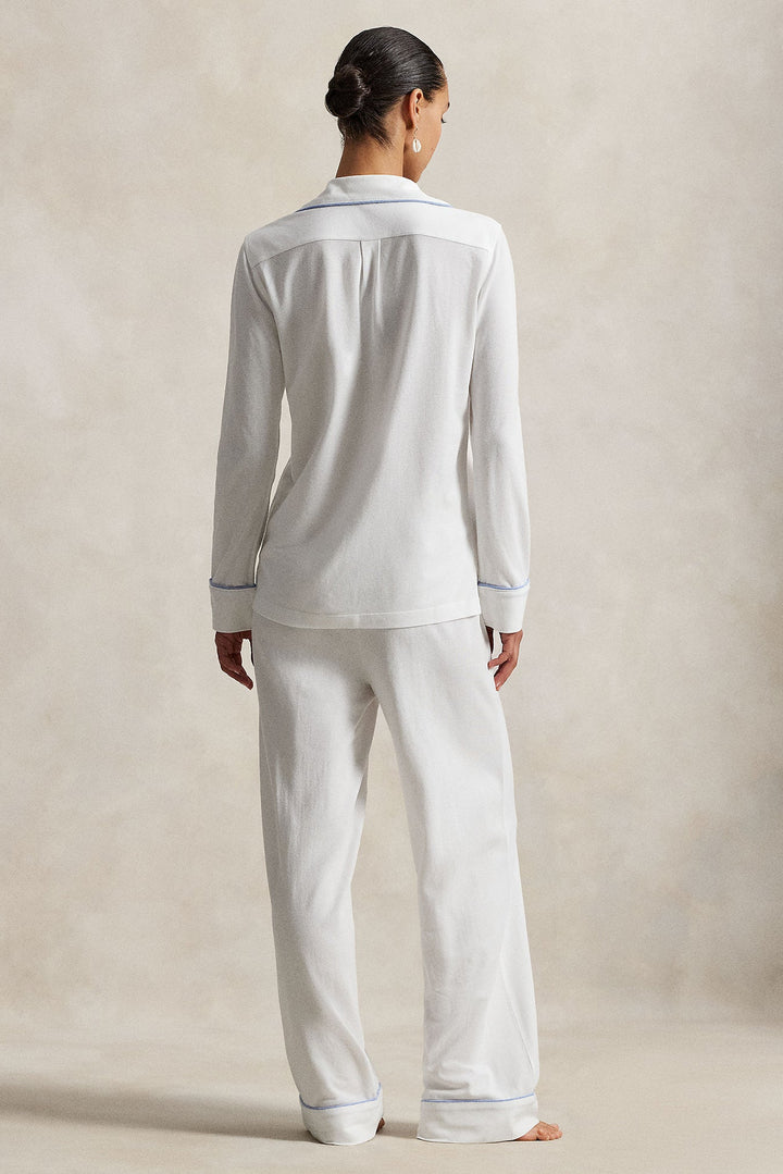 A woman viewed from the back wearing a white long-sleeve pajama shirt and matching wide-leg pajama pants with thin blue piping trim, set against a neutral beige studio backdrop.