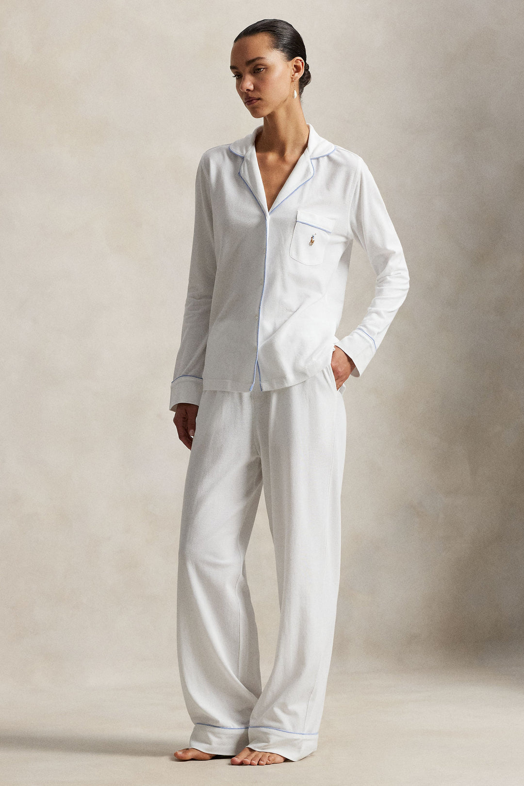 A woman modeling a white long-sleeve pajama set with light blue piping, standing against a textured beige studio backdrop.
