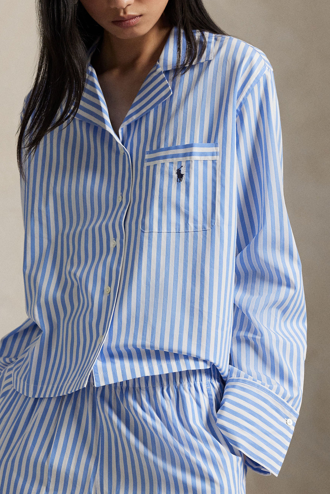 A close-up of a model wearing a blue and white vertically striped long-sleeve button-up shirt with a chest pocket featuring a small embroidered polo player logo, paired with matching striped bottoms.