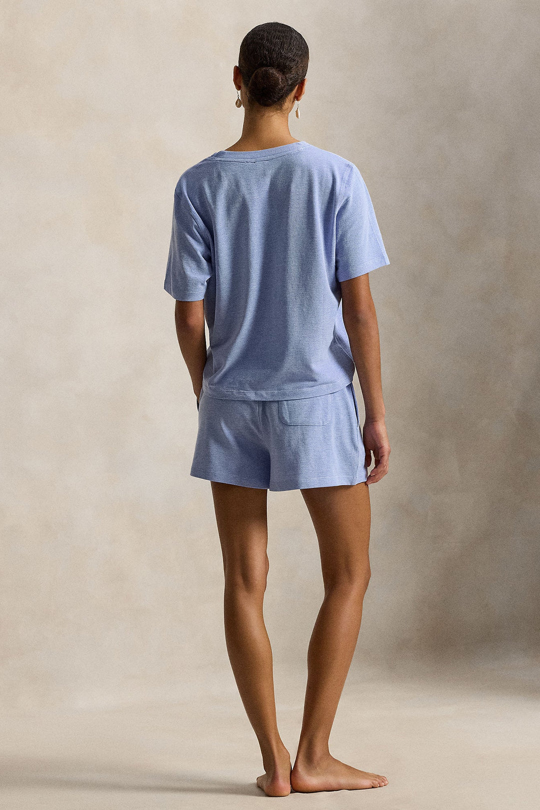 A woman stands barefoot facing away from the camera, wearing a light blue short-sleeve t-shirt and matching shorts set against a textured beige studio backdrop.