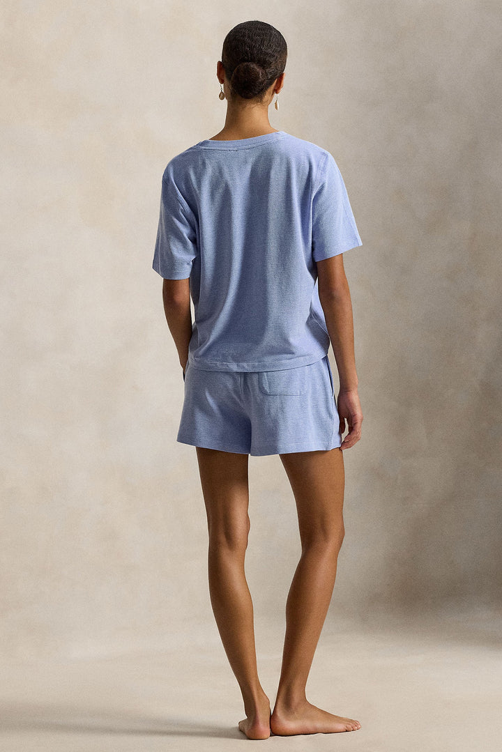 A woman stands barefoot facing away from the camera, wearing a light blue short-sleeve t-shirt and matching shorts set against a textured beige studio backdrop.