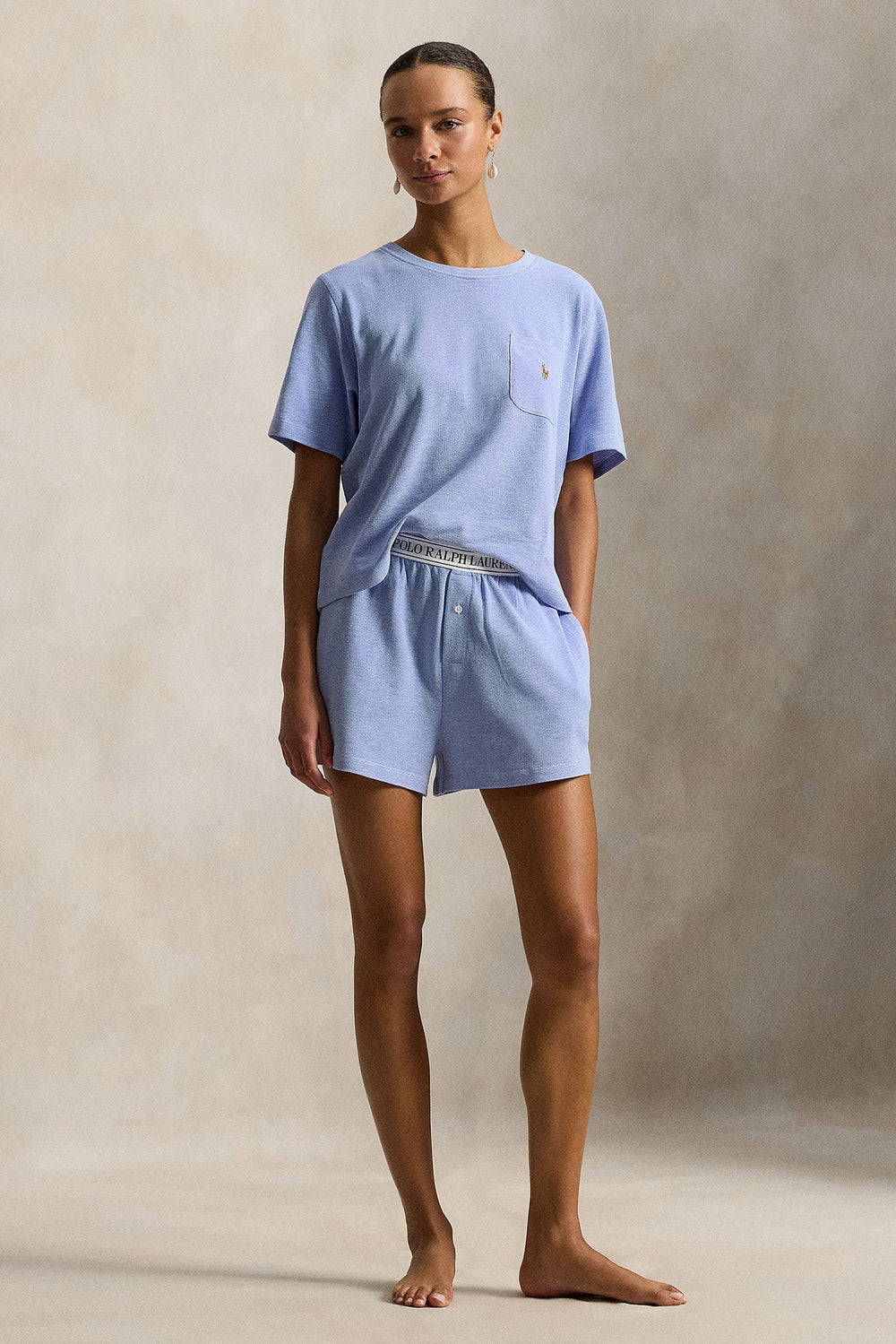 A model stands barefoot against a textured beige studio backdrop wearing a light blue short-sleeve crewneck t-shirt and matching light blue shorts. The shorts feature a waistband with the word 'Polo' printed on it.