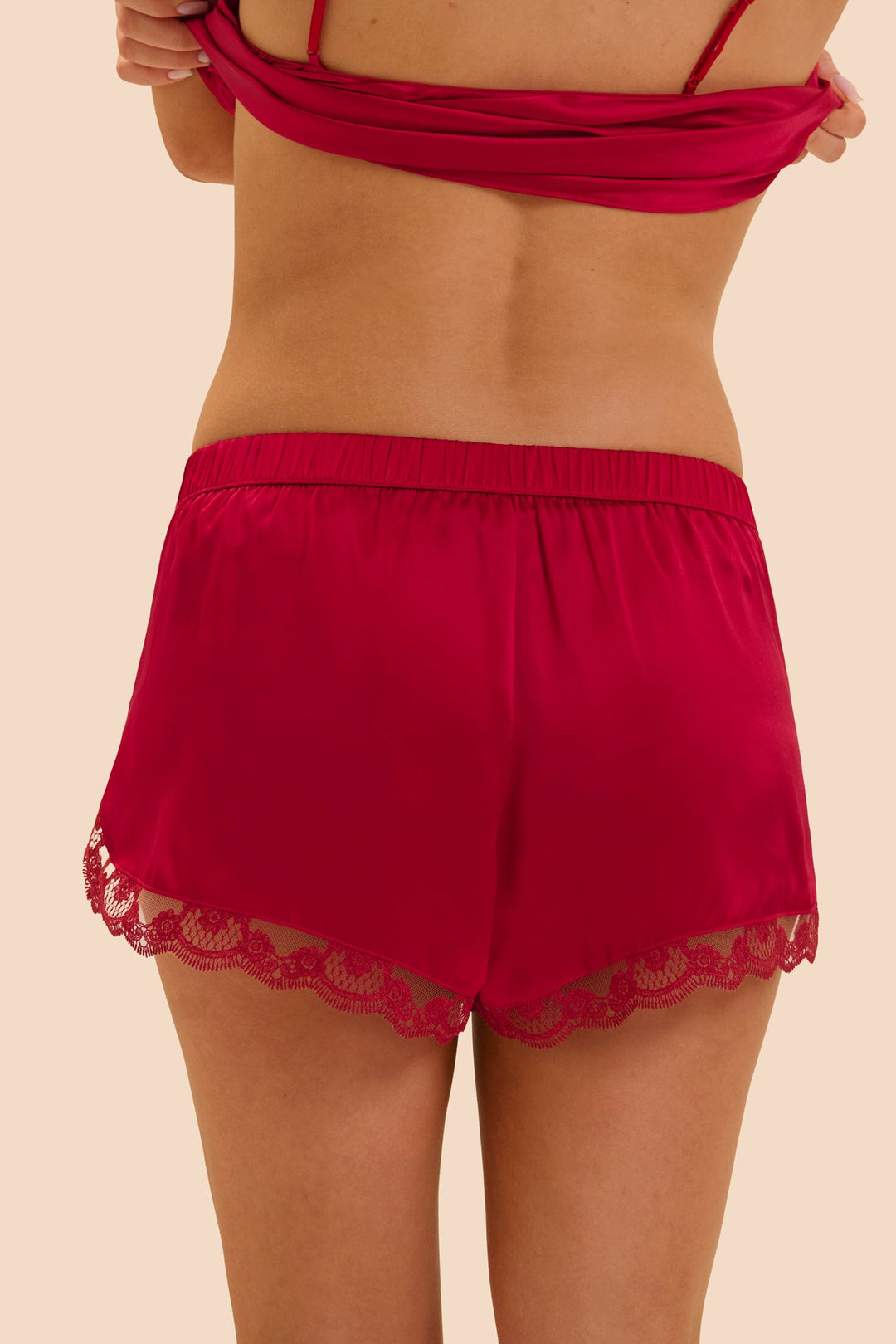 A model from the back wearing bright red satin shorts with a lace hem and a matching red halter-style top, against a solid beige background.