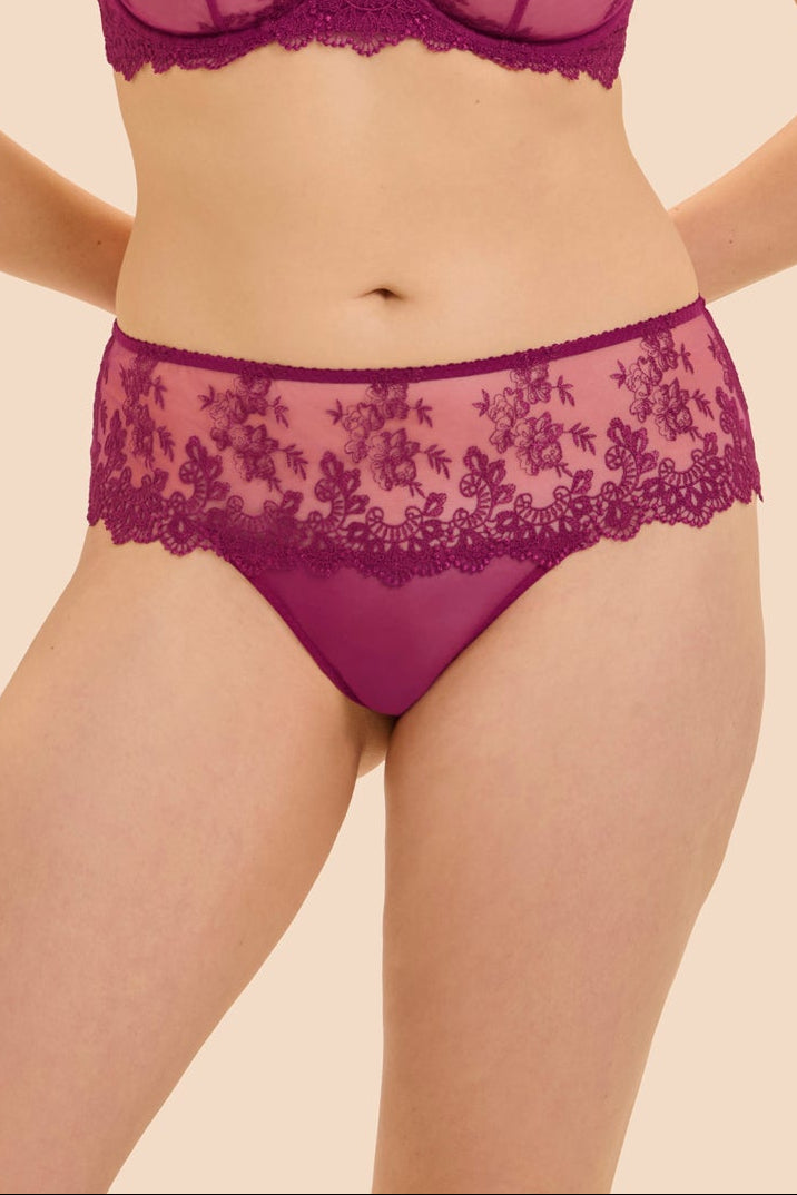 A woman modeling a matching magenta lace bra and panty set against a solid beige background.
