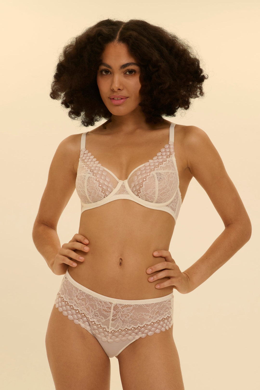 Woman wearing a white lace bra and panties set against a beige background. Pretty bridal, spring French lingerie set.