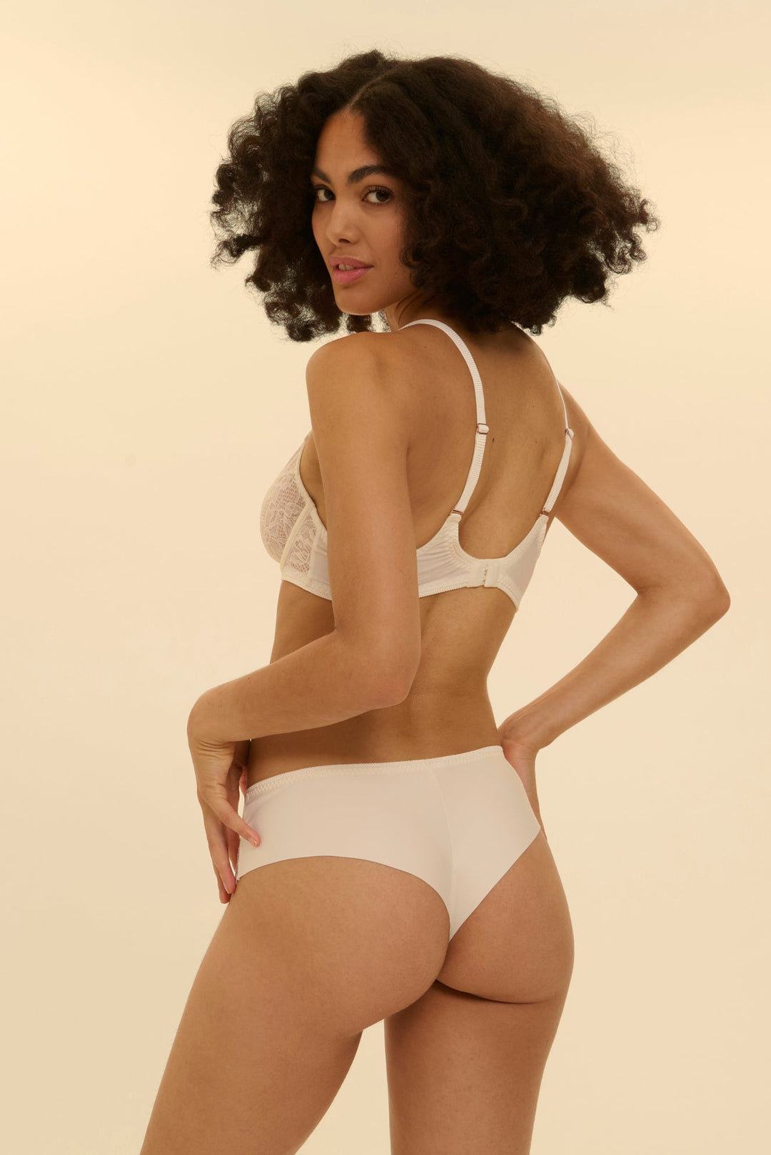 Woman wearing a white lace bra and panties set against a beige background. Pretty bridal, spring French lingerie set.