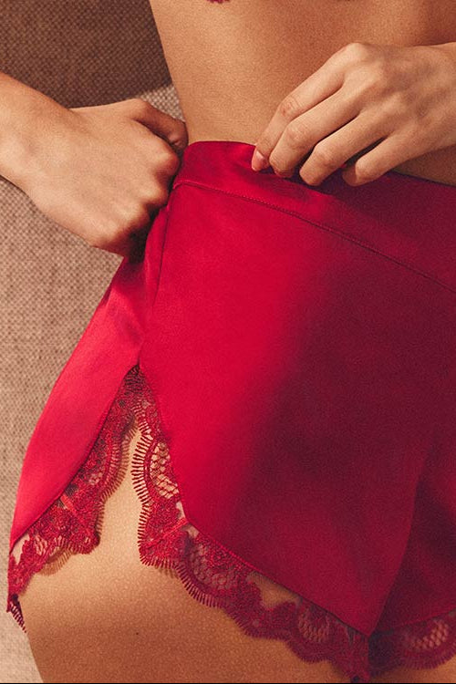 A close-up of a model wearing bright red silk satin shorts with red lace trim, paired with a matching red lace bra. The model is shown from the chest down, adjusting the waistband of the shorts against a neutral textured background.