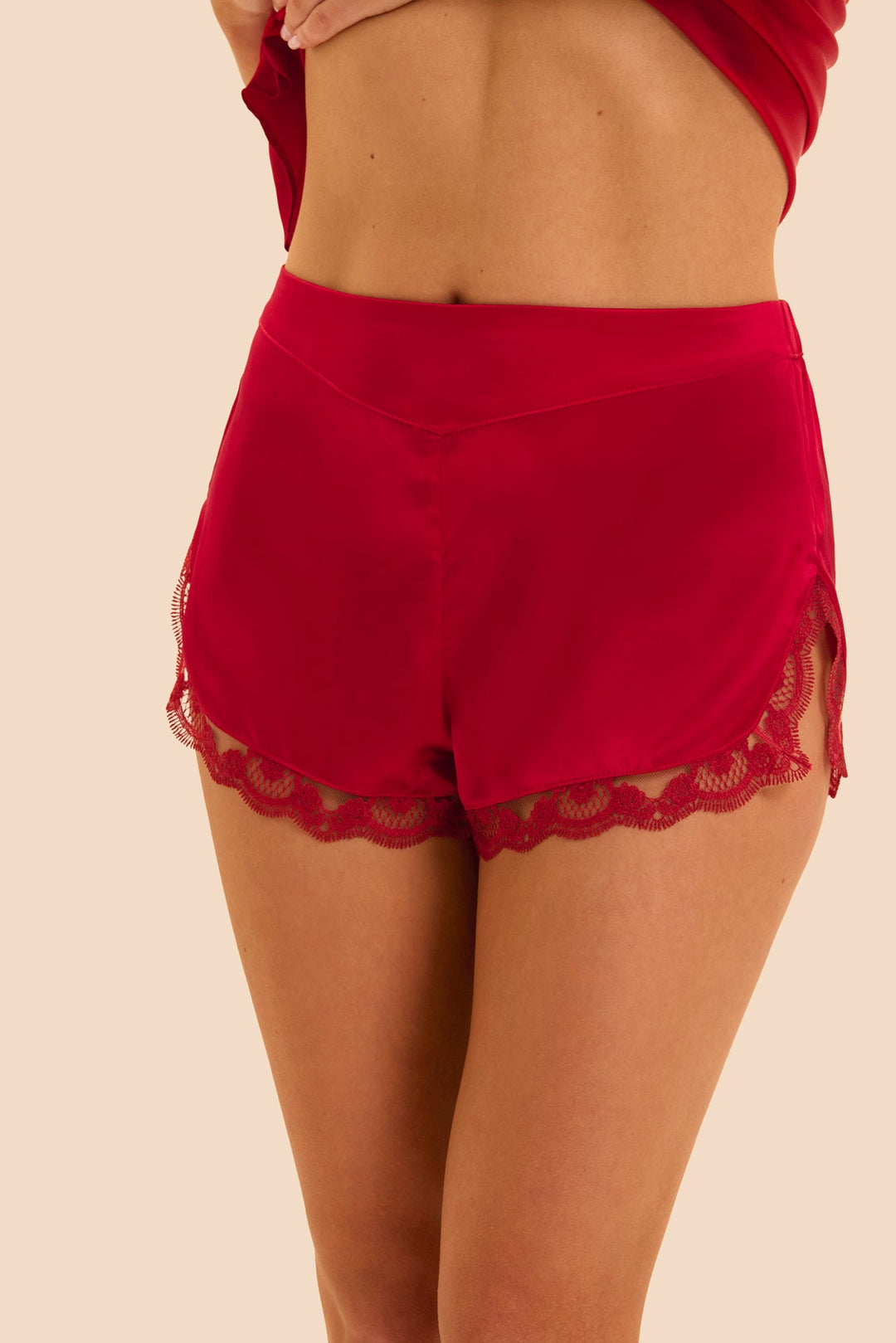 A close-up of a model wearing bright red high-waisted shorts with a scalloped lace hem, shown from the back against a solid beige background.