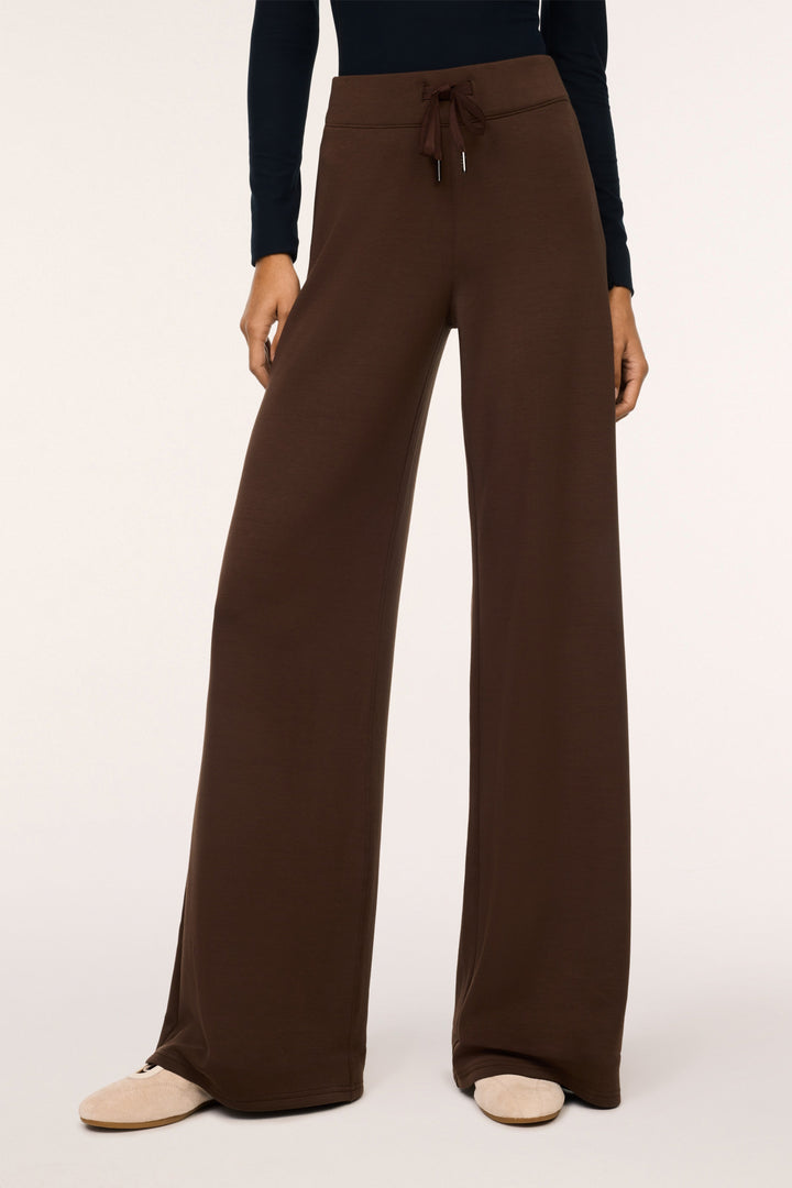 A model wearing dark brown wide-leg drawstring pants, a dark navy long-sleeve top, and beige flat shoes, photographed against a clean off-white studio background.