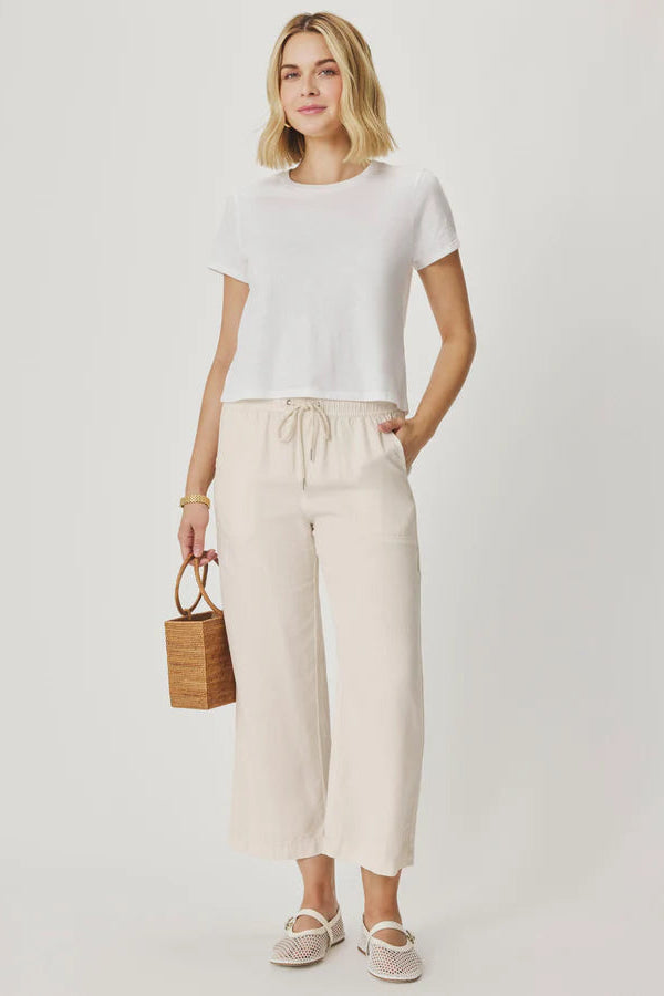 Woman wearing a white top and beige pants holding a woven bag on a plain background