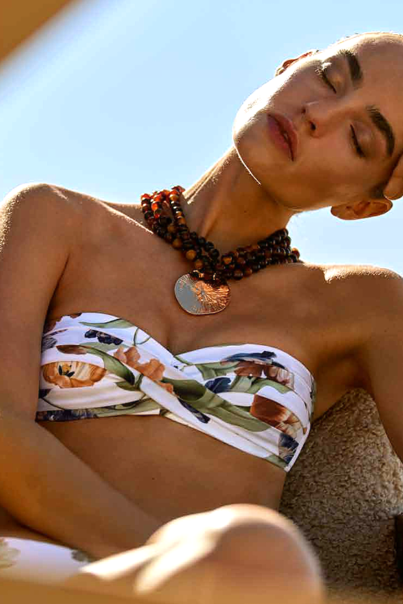 A woman outdoors wearing a white strapless bikini top with a botanical and animal print, paired with layered dark brown beaded necklaces.