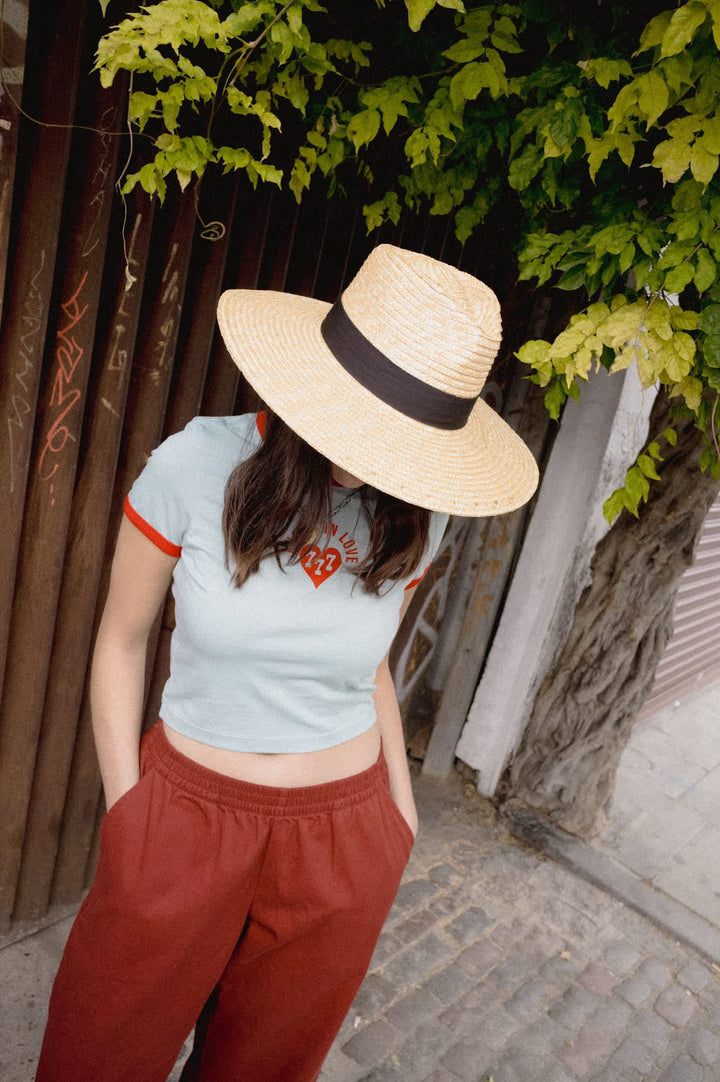A person wearing a wide-brim straw boater hat with a black band, a light blue cropped t-shirt with red trim and a small red heart graphic, and rust-colored pants, standing outdoors under green leaves.